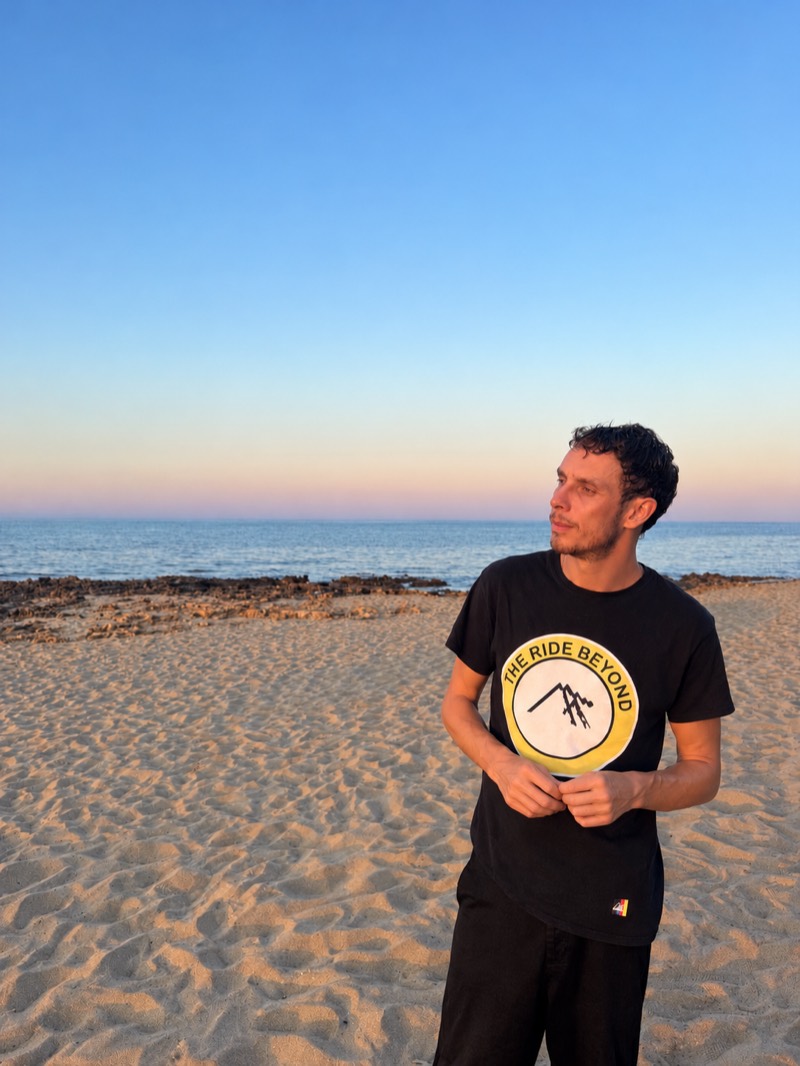 Liam Desroy on a sandy beach at sunset, looking out to sea, hands clasped in front of him